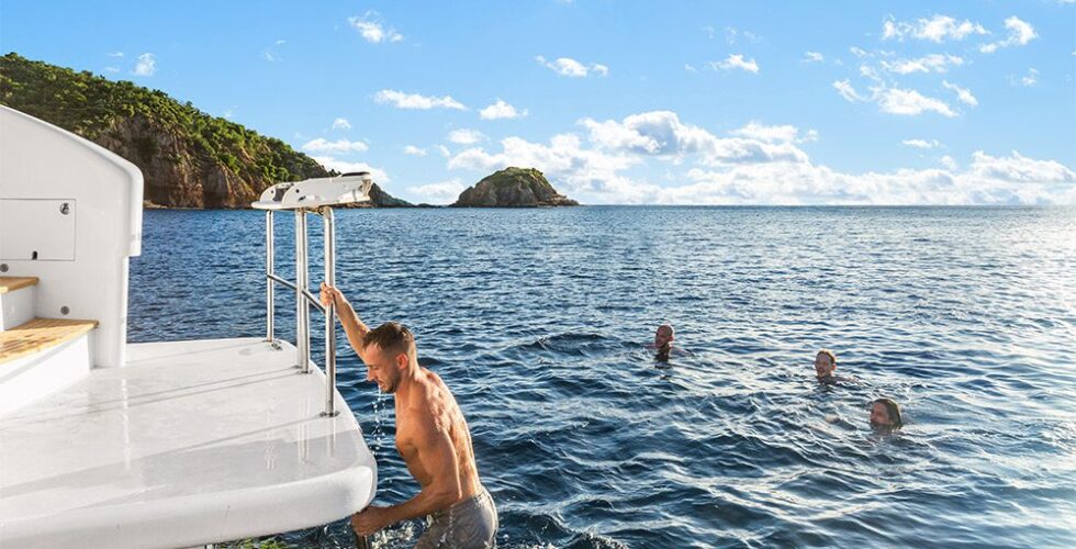 A man climbing onto a luxury power catamaran charter ladder with others swimming in the sea near a coastal landscape.